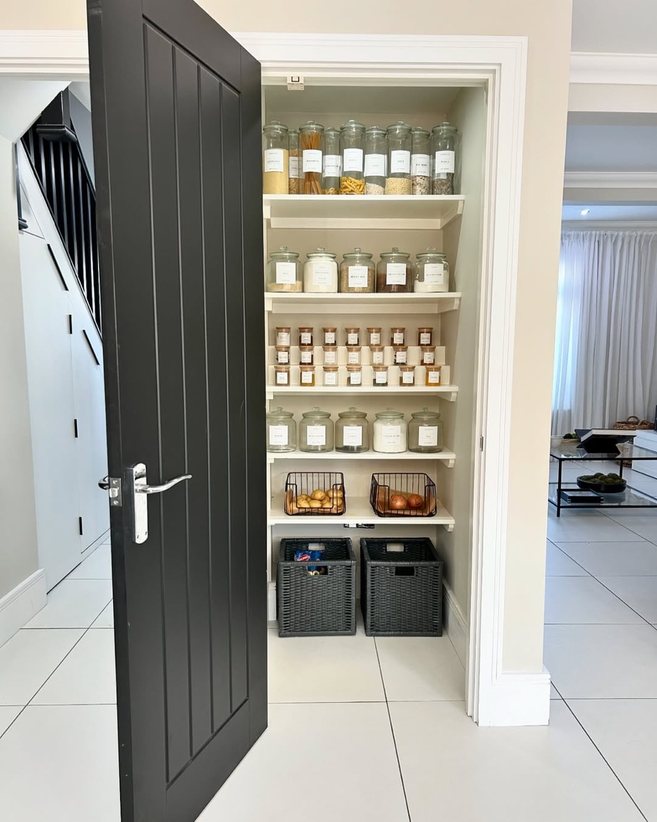 Compact reach-in pantry with uniform glass jars, labeled spices, and wire baskets behind a dark door, Roslyn Heights Long Island